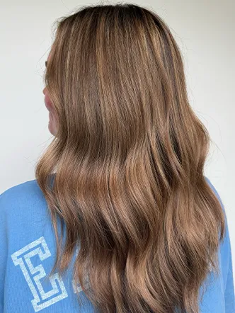Person with long, wavy, light brown hair facing away from the camera, wearing a light blue top with white lettering on the back—a look that could be achieved at the best balayage and highlights in Spring Hill TN. | Salon Blonde in Columbia, TN, Spring Hill, TN, Franklin, TN