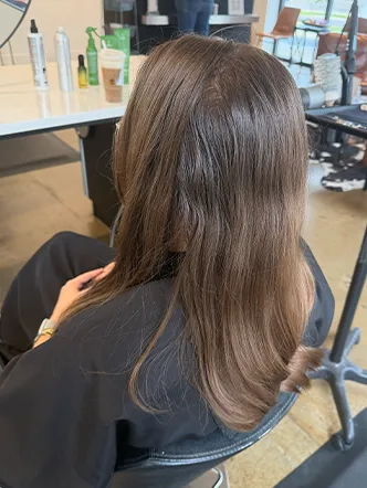 Person with straight, medium-length brown hair sits in a salon chair, wearing a black cape. Hair products and a drink are visible on the table in the background, creating a welcoming atmosphere at the best hair salon in Franklin TN. | Salon Blonde in Columbia, TN, Spring Hill, TN, Franklin, TN