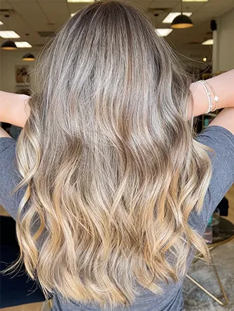 Person with long, wavy, ash blonde hair viewed from the back, standing indoors under ceiling lights at the best hair salon in Franklin TN. | Salon Blonde in Columbia, TN, Spring Hill, TN, Franklin, TN