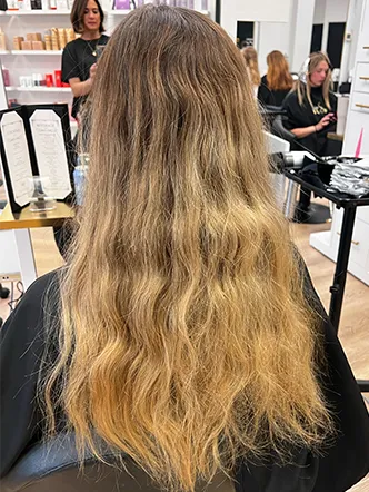 Person with long, wavy, blonde and brown hair sits in a salon chair, facing away from the camera. Hair products and salon staff are visible in the background, reflecting the expertise behind some of the best balayage and highlights in Spring Hill TN. | Salon Blonde in Columbia, TN, Spring Hill, TN, Franklin, TN