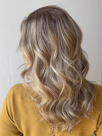Person with long, wavy blonde hair—styled to perfection by the best hair salon in Franklin TN—wearing a mustard yellow top, shown from the back against a plain background. | Salon Blonde in Columbia, TN, Spring Hill, TN, Franklin, TN