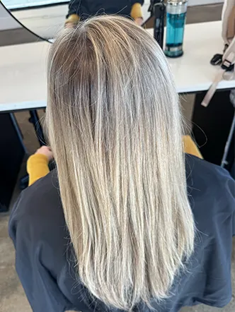 Person with straight, shoulder-length blonde hair sits in front of a salon mirror at the best hair salon in Franklin TN, wearing a dark cape and yellow sleeves visible, with premium hair products on the counter. | Salon Blonde in Columbia, TN, Spring Hill, TN, Franklin, TN