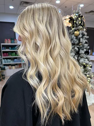 A person with long, wavy, blonde hair sits in a salon—widely known as the best hair salon in Franklin TN—with shelves of products and a decorated Christmas tree visible in the background. | Salon Blonde in Columbia, TN, Spring Hill, TN, Franklin, TN