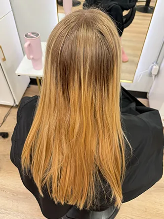 Person with long, straight hair that transitions from darker blonde at the roots to lighter golden blonde at the ends, seated in a salon chair—showcasing results from the best balayage and highlights in Spring Hill TN. | Salon Blonde in Columbia, TN, Spring Hill, TN, Franklin, TN