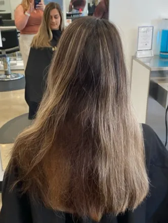 A person with shoulder-length, brown hair with lighter highlights sits in a salon chair facing a mirror, wearing a black cape, ready to experience the best balayage and highlights in Spring Hill TN. | Salon Blonde in Columbia, TN, Spring Hill, TN, Franklin, TN