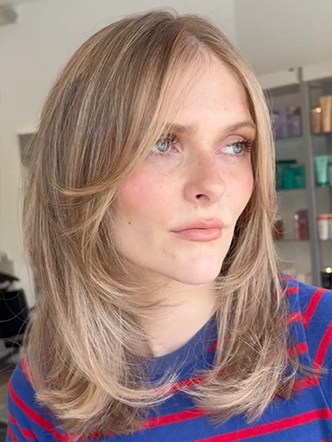 A person with shoulder-length, layered blonde hair—showcasing the best balayage and highlights in Spring Hill TN—wearing a blue and red striped shirt and looking off to the side in a well-lit room. | Salon Blonde in Columbia, TN, Spring Hill, TN, Franklin, TN
