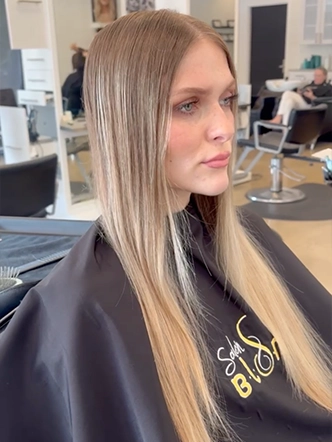 A woman with straight, blonde hair sits in a salon chair wearing a black cape. Salon equipment and mirrors are visible in the background, reflecting the modern ambiance of the best hair salon in Franklin TN. | Salon Blonde in Columbia, TN, Spring Hill, TN, Franklin, TN