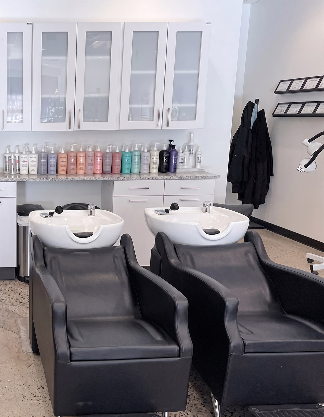 Two black salon shampoo chairs and sinks face forward, with shelves of hair products, cabinets, and hanging robes visible in the background of a modern salon setting—an inviting space that exemplifies the best hair salon in Franklin TN. | Salon Blonde in Columbia, TN, Spring Hill, TN, Franklin, TN