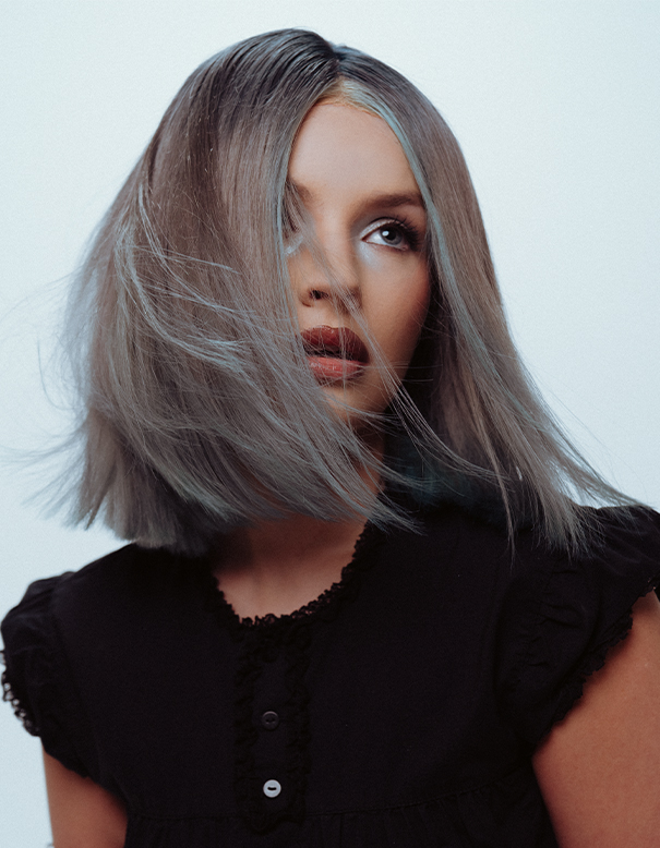 A person with straight, shoulder-length silver hair and blue highlights looks upward. Showcasing one of the best balayage and highlights in Spring Hill TN, they wear a black, short-sleeved top with buttons and lace trim. | Salon Blonde in Columbia, TN, Spring Hill, TN, Franklin, TN