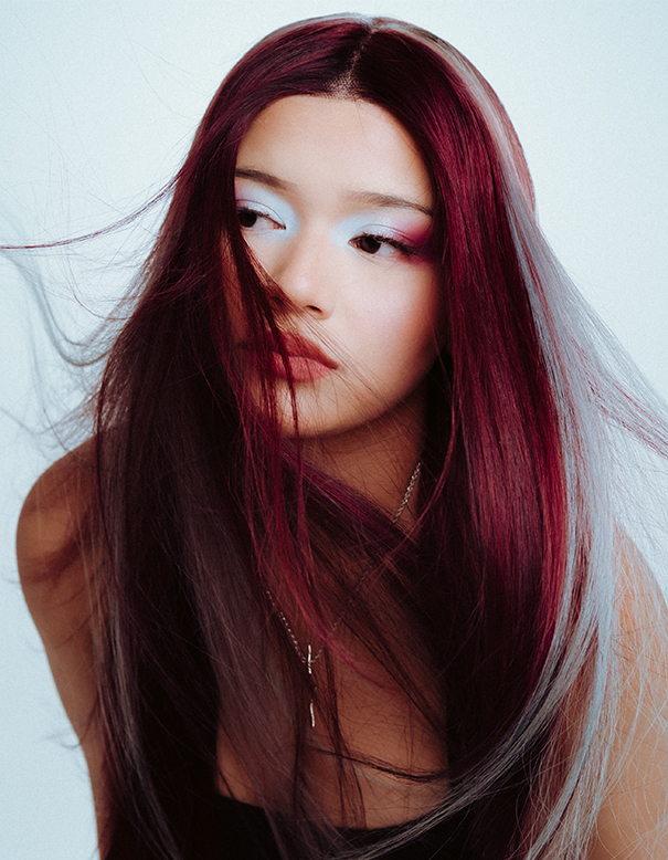 A person with long, straight, red and silver hair—styled as if by the best hair salon in Franklin TN—looks to the side. They have light makeup and wear a black top and silver necklace against a plain background. | Salon Blonde in Columbia, TN, Spring Hill, TN, Franklin, TN