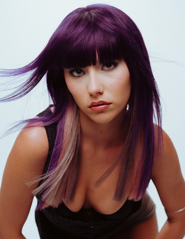 Woman with straight, purple hair and blunt bangs looks at the camera with a neutral expression. She is wearing a black top with a low neckline against a plain background, showcasing vibrant color that could only be achieved at the best hair salon in Franklin TN or by using the best hair extensions in Franklin TN. | Salon Blonde in Columbia, TN, Spring Hill, TN, Franklin, TN
