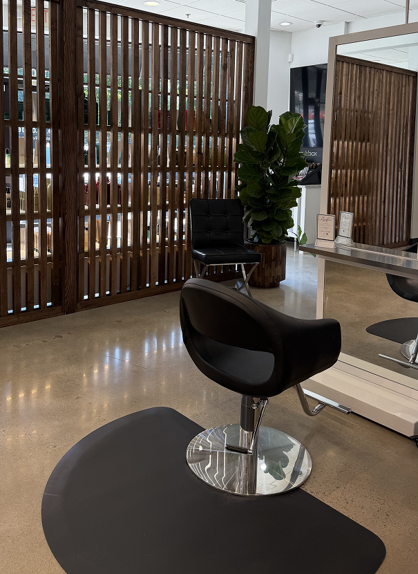 A modern salon features a black styling chair, a mirror station, a potted plant, and wooden slat partitions on a polished concrete floor. | Salon Blonde in Columbia, TN, Spring Hill, TN, Franklin, TN