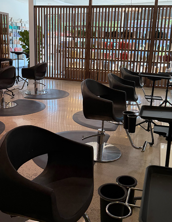 Modern hair salon interior with black styling chairs, round floor mats, workstations, and wooden partitions; shelves with products visible in the background. | Salon Blonde in Columbia, TN, Spring Hill, TN, Franklin, TN