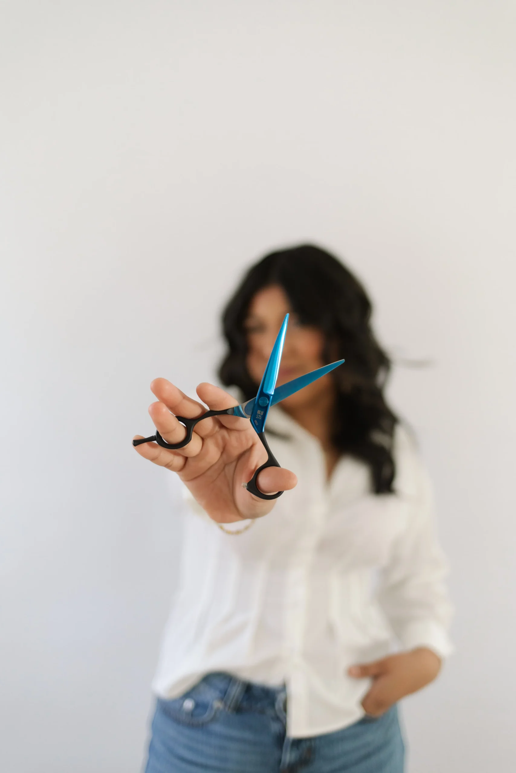 Person in a white shirt holds blue scissors toward the camera, with face out of focus against a plain white background—capturing the modern, fresh vibe you’ll find at the salon known for the best balayage and highlights in Spring Hill TN. | Salon Blonde in Columbia, TN, Spring Hill, TN, Franklin, TN