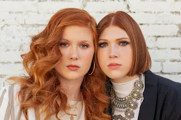Two women with red hair pose together in front of a white brick wall, one with long curly hair and the other with a straight bob, both wearing neutral-toned clothing—showcasing styles created by the best hair salon in Franklin TN. | Salon Blonde in Columbia, TN, Spring Hill, TN, Franklin, TN