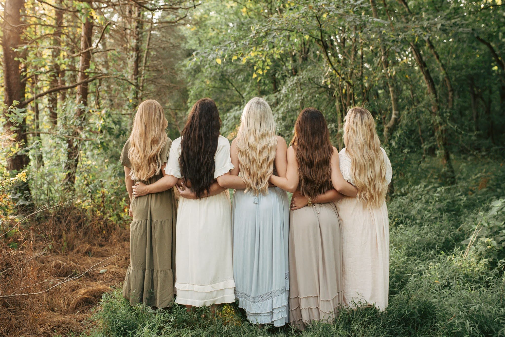 Five women with long hair stand side by side in a green forest, arms around each other, wearing light-colored dresses, seen from behind—each showcasing the luxurious results you can achieve at the best hair salon in Franklin TN. | Salon Blonde in Columbia, TN, Spring Hill, TN, Franklin, TN
